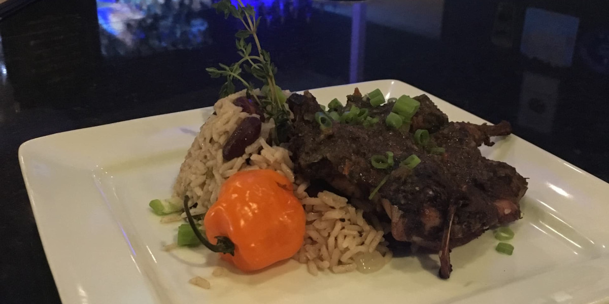 In a dimly lit restaurant at Spare Time Texas, enjoy a plate of jerk chicken served with rice and peas, garnished with fresh thyme and chopped green onions. A whole orange habanero pepper graces the white square plate—a perfect meal after an exciting round of bowling.