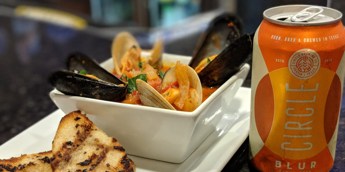 A bowl of mussels and clams in a tomato-based sauce sits on a white plate, accompanied by slices of grilled bread. Beside the plate is a can of Circle Blur beer with an orange label, perfect after a day at Spare Time Texas, where family fun and bowling await.