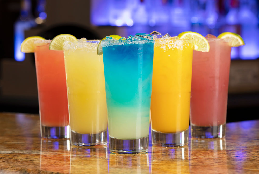 Five colorful cocktails grace the wooden bar at Spare Time Texas, each glass filled with ice and garnished with lime wedges. In shades of pink, yellow, blue, and orange, they create a vibrant display perfect for unwinding after a thrilling game of bowling.
