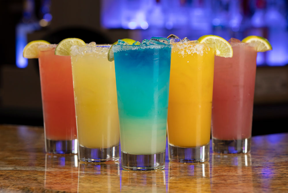 A row of colorful cocktails on a bar counter at Spare Time Texas invites you to unwind after a day of strikes and spares. The drinks, in tall glasses with salted rims and lime wedges, feature pink, yellow, blue, orange hues. The softly lit blue background adds to the family fun center's vibrant atmosphere.
