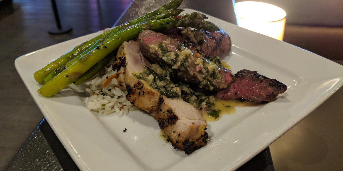 A plate with roasted chicken and steak slices garnished with herbs, served over rice, is reminiscent of a meal enjoyed at Spare Time Texas. Grilled asparagus spears are placed on top under the soft glow of a candle, echoing the warm ambiance of this beloved family fun center.