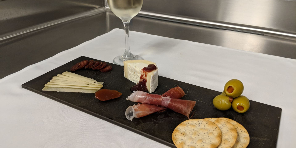A slate cheese board on a white cloth features slices of manchego, brie with jam, cured meats, green olives, and crackers. A tall wine glass filled with white wine stands in the background—perfect for savoring during a relaxing evening at Spare Time Texas after a fun game of bowling.