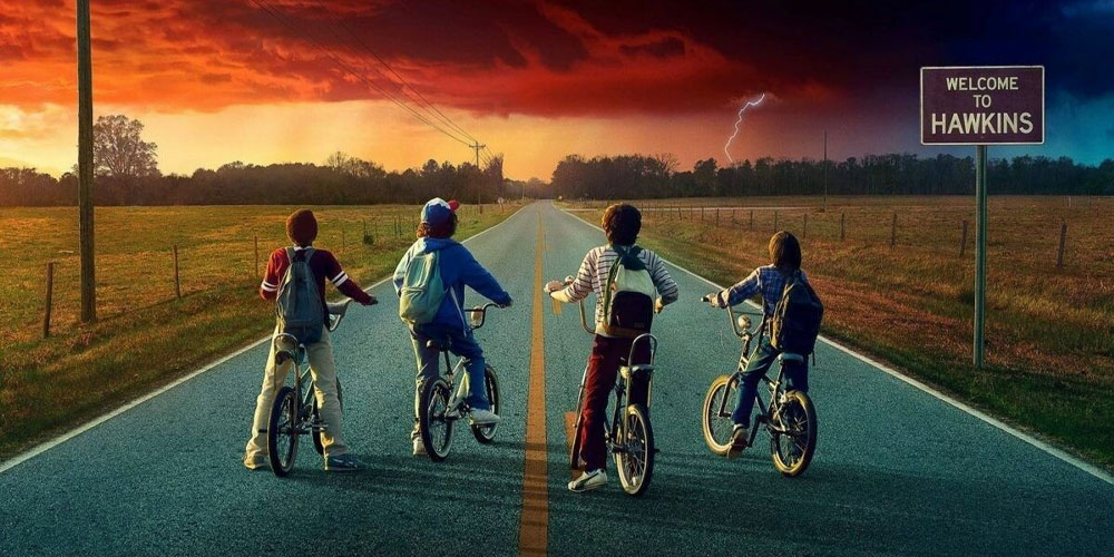 Four kids on bikes are facing a stormy, red sky as they ride down a rural road. A sign reads "Welcome to Hawkins." Lightning strikes in the distance, casting an eerie glow reminiscent of adventures at Spare Time Texas, where every turn hints at mystery.